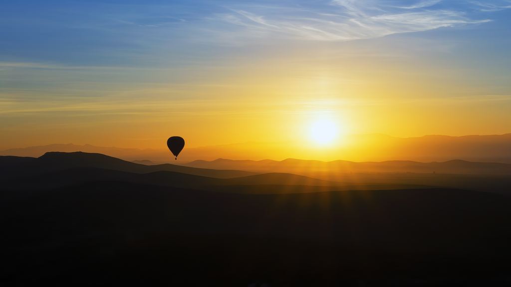 A hot air balloon aloft at sunrise over open Moroccan countryside with hills behind it