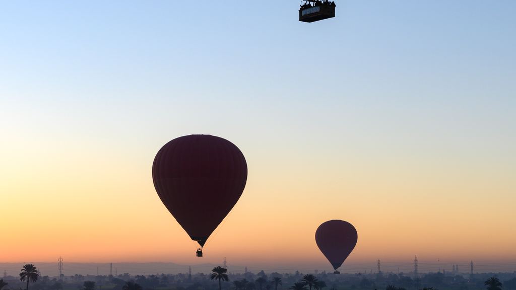 Hot air balloons at sunrise above irrigated fields, with the red cliffs of the Luxor west bank behind them.