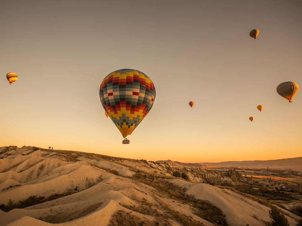 A hot air balloon over the eroded valleys of Cappadocia at sunrise, with more balloons scattered across the sky behind it.