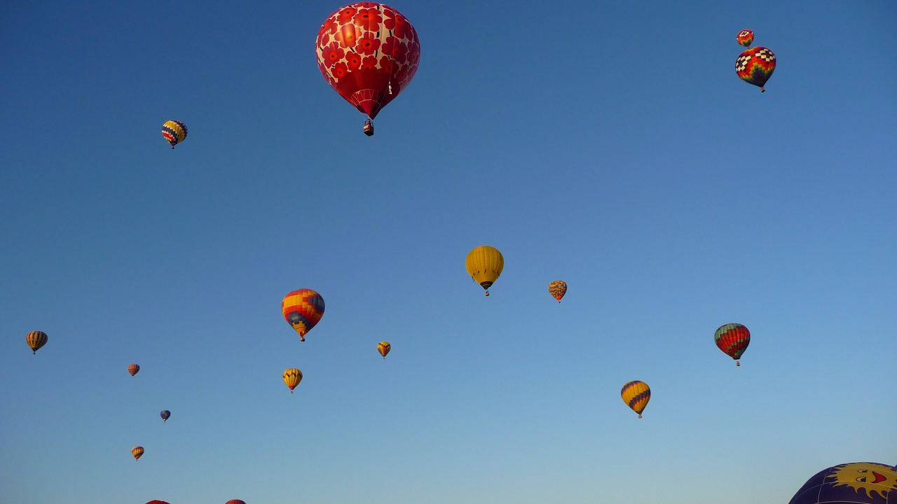People walking among hot air balloons on the launch field as dozens more fill the sky at the Albuquerque Balloon Fiesta