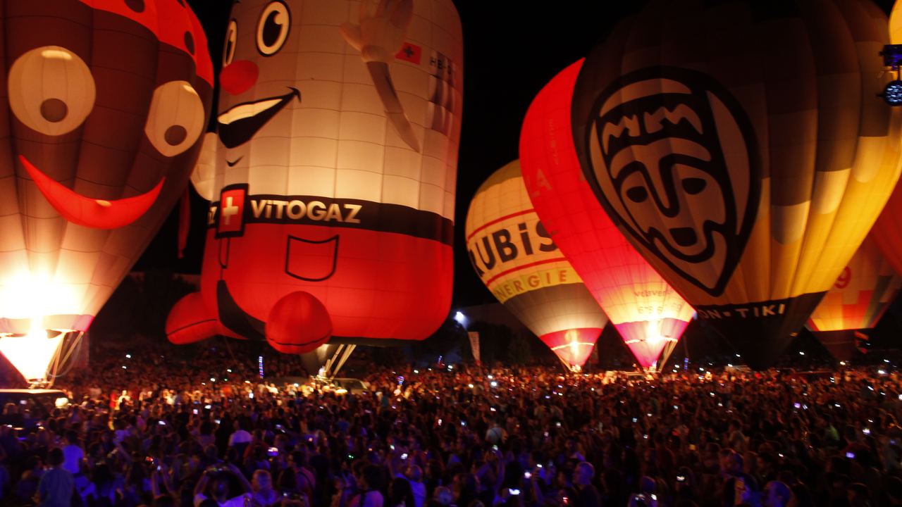 Hot air balloons tethered on the ground at night, envelopes lit from inside by their burners during a night glow