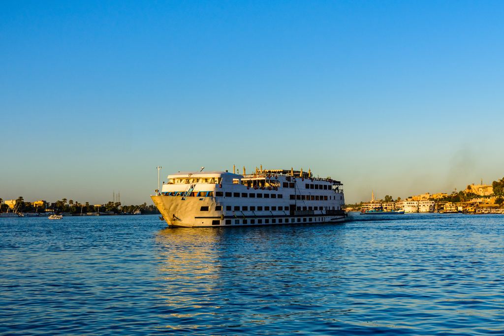 A white multi-deck Nile cruise boat under way on the river in late afternoon light, with low buildings along the far bank.
