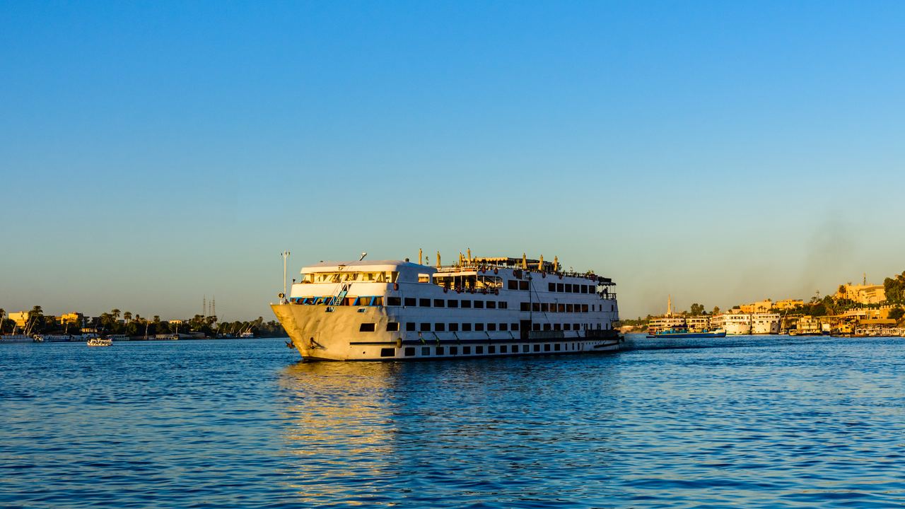 A white multi-deck Nile cruise boat under way in late afternoon light, with low buildings along the far bank